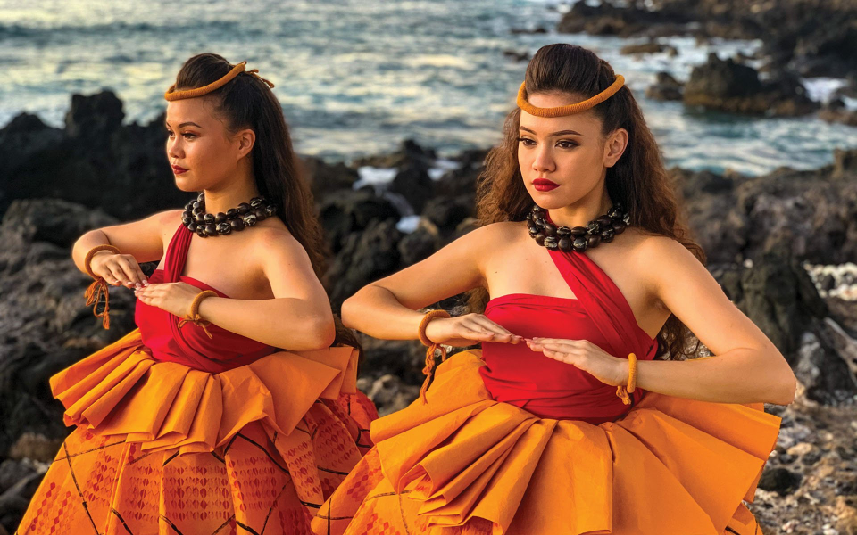 Luau At Mauna Kea Beach Hotel: two young women in read and orange traditional dress dancing hula at the seashore